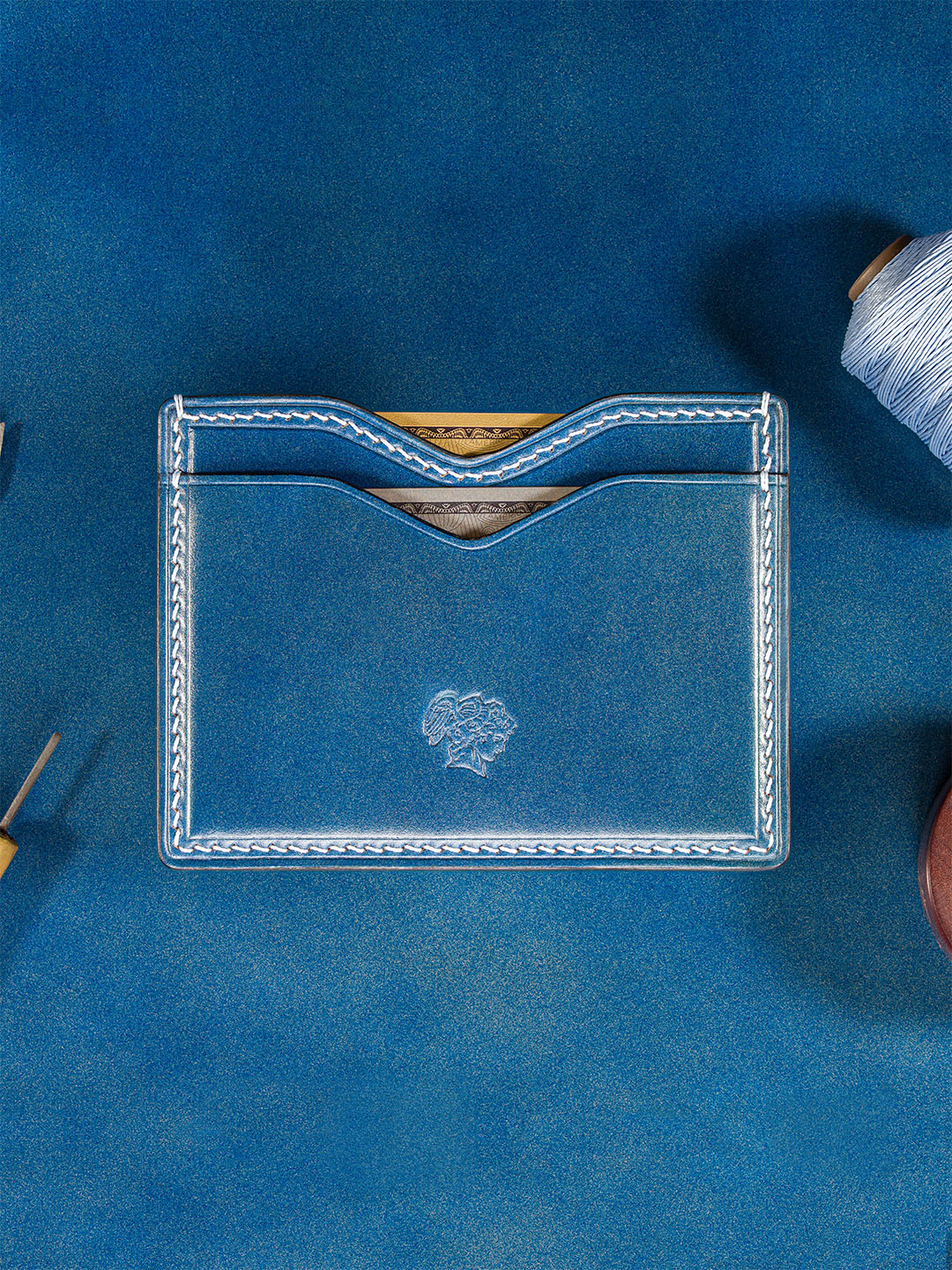 Berling Heirloom card holder in vivid blue shell cordovan, with light blue stitching, an embossed logo and two cards in its curved pockets. Shown on a blue surface beside leatherworking tools and a spool of thread.