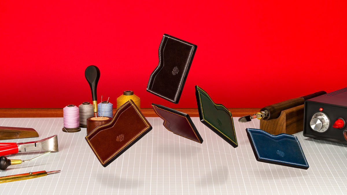 Handstitched shell cordovan card holders in brown, burgundy, green and blue floating above a leatherworker's cutting mat